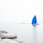 A small white rowing boat moored on archipelago rocks while a sailboat with a bold blue sail glides past on open water.